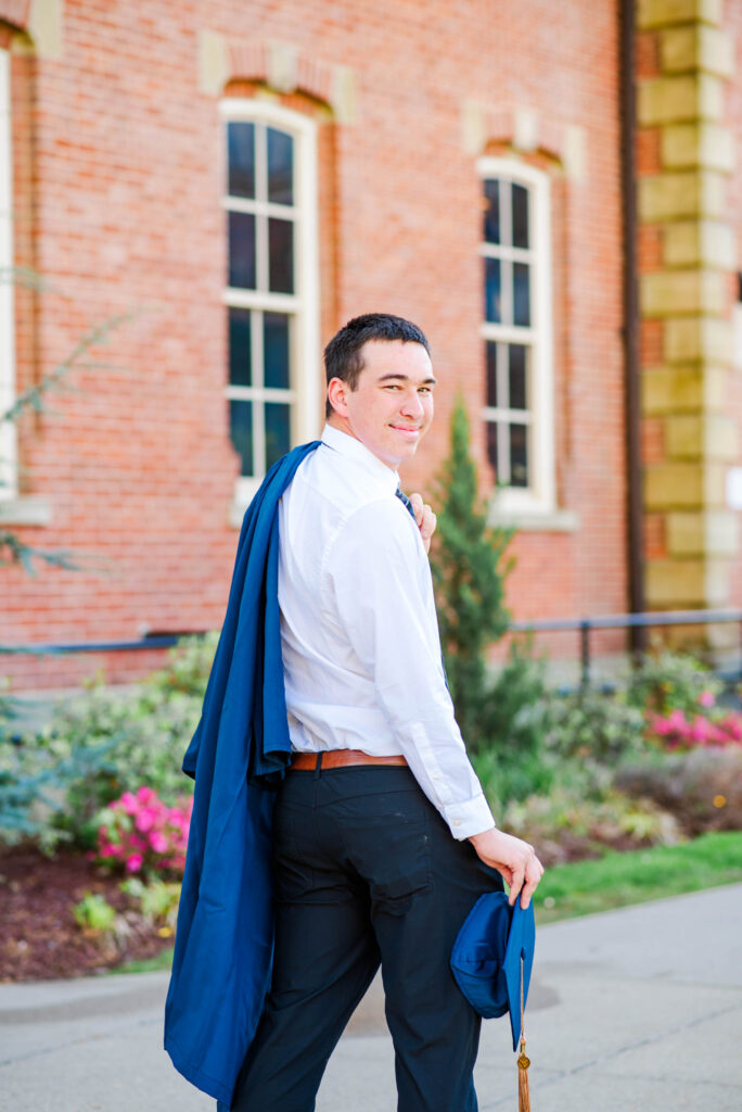 graduationphotoideasfor guys040 carrie white photography young man in a white shirt with dark pants a blue graduation gown draped over his shoulder standing outdoors in front of a brick campus building and smiling