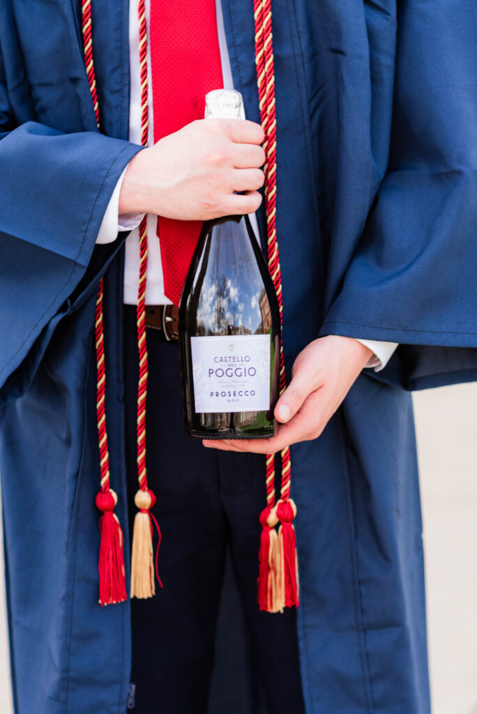 graduationphotoideasfor guys041 carrie white photography graduate in a blue gown holding a castello del poggio prosecco bottle with red tassels around the bottle