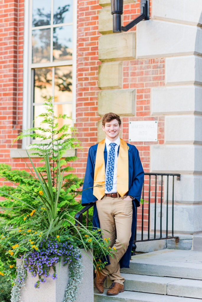 graduationphotoideasfor guys050 carrie white photography graduation photo ideas for guys shows a grad in blue gown and gold stole stands on stone steps outside a brick building with greenery in the foreground smiling at the camera