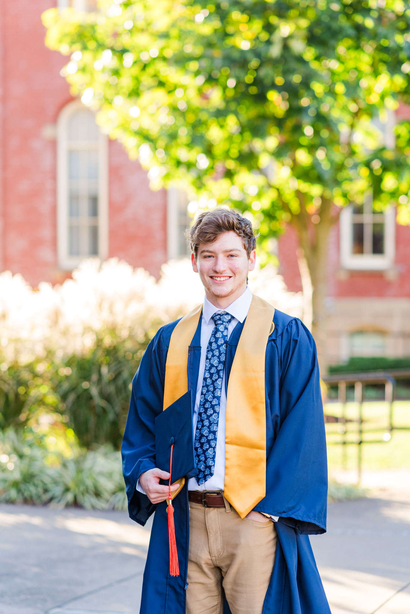 male graduate in a navy cap and gown with a gold stole smiling outdoors on campus