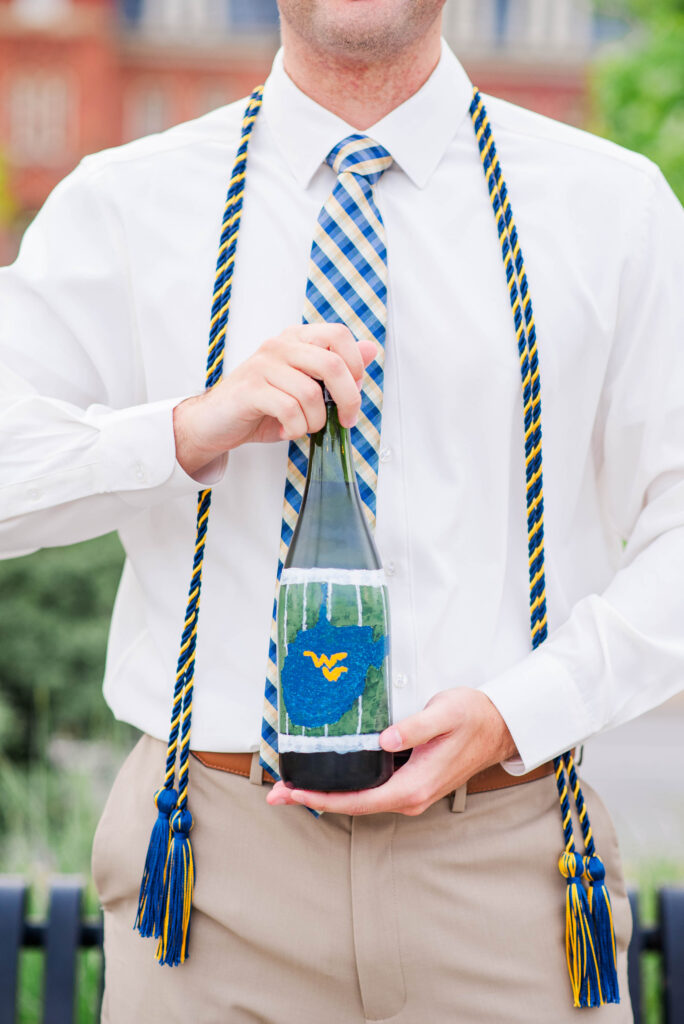 graduationphotoideasfor guys088 carrie white photography man in a white dress shirt and blue plaid tie holding a champagne bottle outdoors with blue and gold graduation cords draped over his shoulders