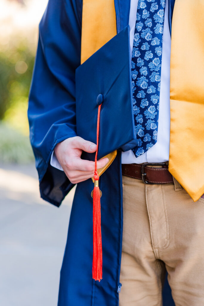 graduationphotoideasfor guys094 carrie white photography graduate in a blue gown with a yellow stole holding a matching cap with a red tassel visible at the front