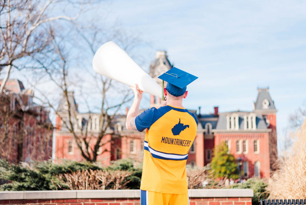 graduationphotoideasfor guys11 carrie white photography back view graduate in blue cap and orange gown raises a white megaphone outdoors'Mountaineers' on gown visible