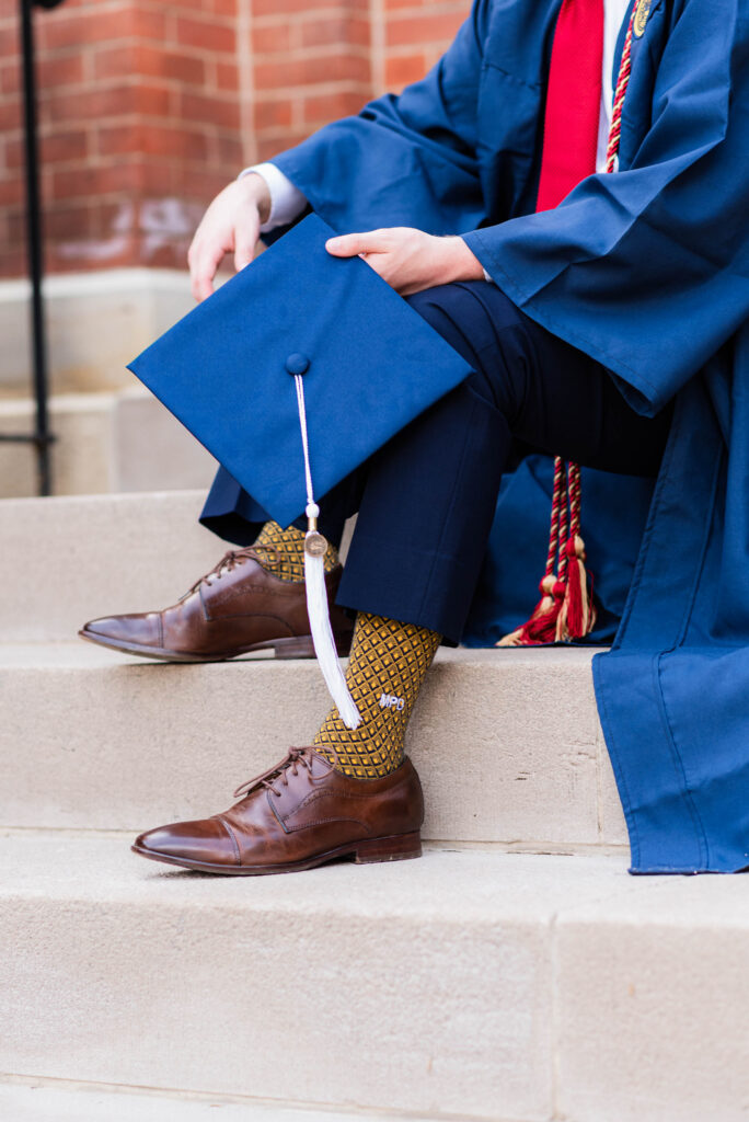 graduationphotoideasfor guys103 carrie white photography graduation photo ideas for guys show graduate in a blue cap and gown sits on concrete steps holding the mortarboard with a white tassel brown dress shoes and patterned socks visible