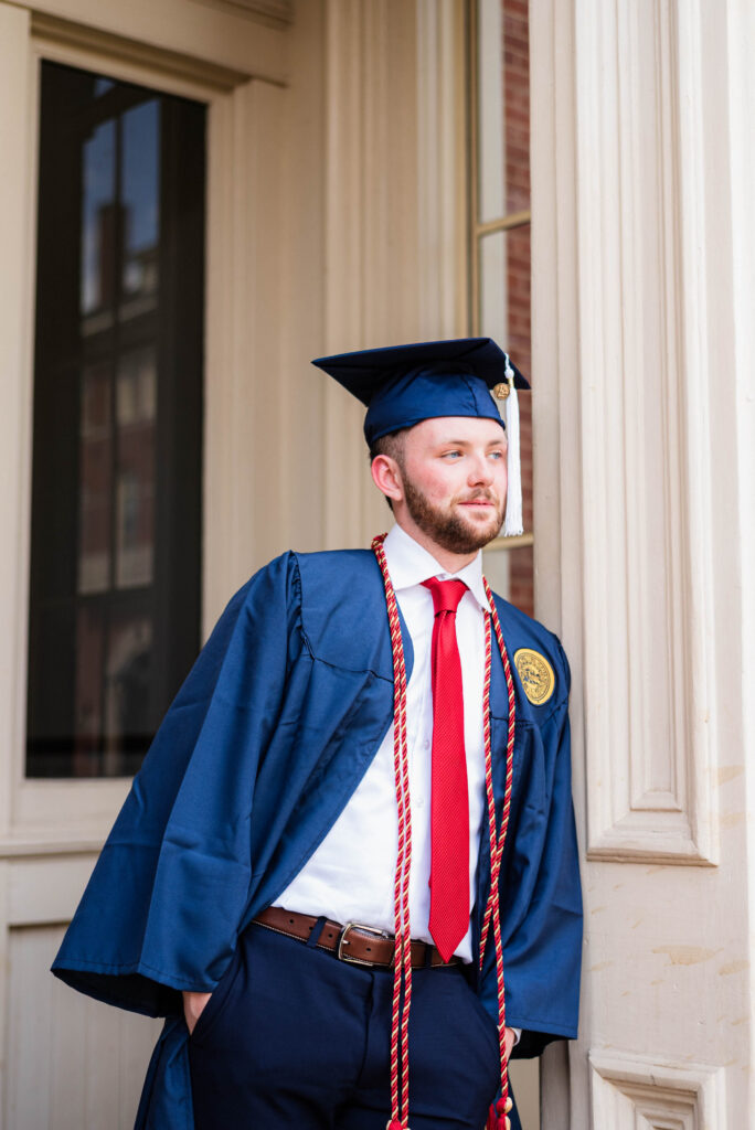 graduationphotoideasfor guys109 carrie white photography male graduate in a blue cap and gown with red cords posed beside a column outside a building
