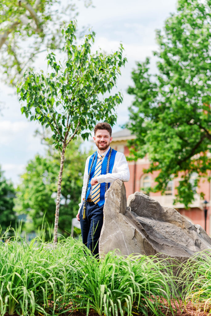 graduationphotoideasfor guys117 carrie white photography smiling man in white shirt with blue graduation stole and cords standing by large rocks in a green courtyard for wvu spring grad photos