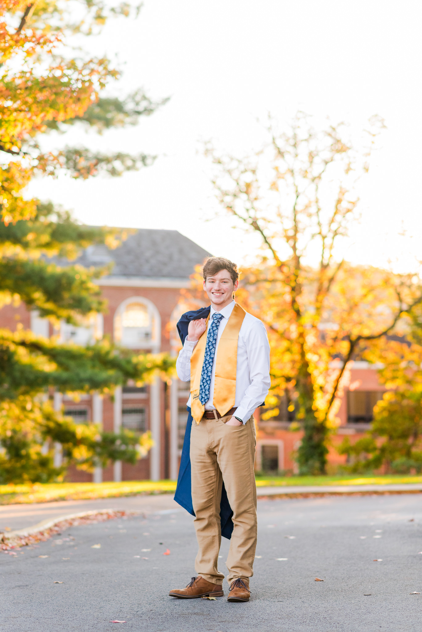 Young graduate standing outdoors in a gown and gold stole on a campus quad surrounded by autumn trees.