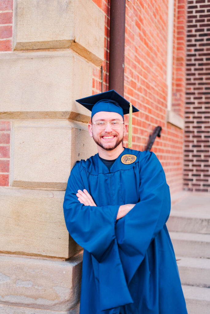 graduationphotoideasfor guys31 carrie white photography graduate in a blue cap and gown leaning against a brick and stone wall arms crossed and smiling outdoors
