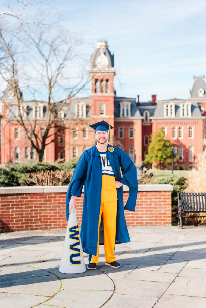 graduationphotoideasfor guys41 carrie white photography graduate in a blue cap and gown on campus holding a white megaphone with blue lettering with red brick buildings in the background