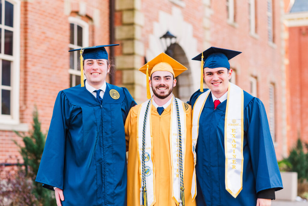 theforencisteam137 carrie white photography three graduates in blue and gold caps and gowns stand together outdoors on campus smiling at the camera