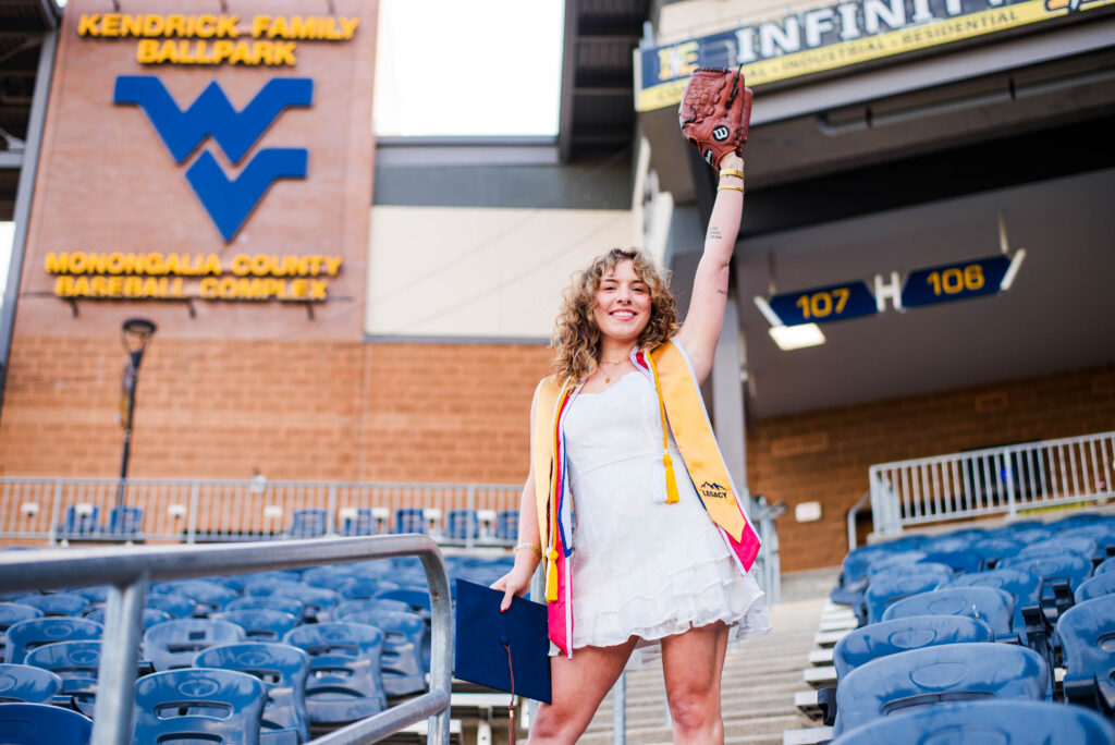 wvubaseballgradphotos03 carrie white photography graduate in a white dress with a yellow graduation stole raises a baseball glove above her head in a stadium with blue seats and signs in the background