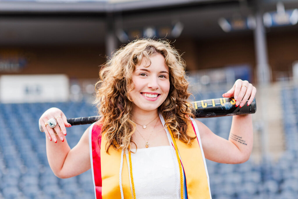 wvubaseballgradphotos11 carrie white photography smiling woman with curly hair holding a bat across her shoulders at a kendrick family ballpark during baseball graduation pictures