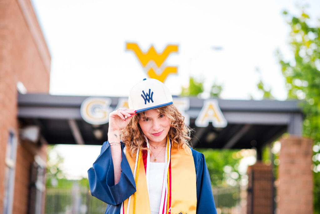 wvubaseballgradphotos10 carrie white photography graduate woman tipping a white cap with blue logo wearing a blue gown and yellow stole outdoors near a campus sign with a wv style logo