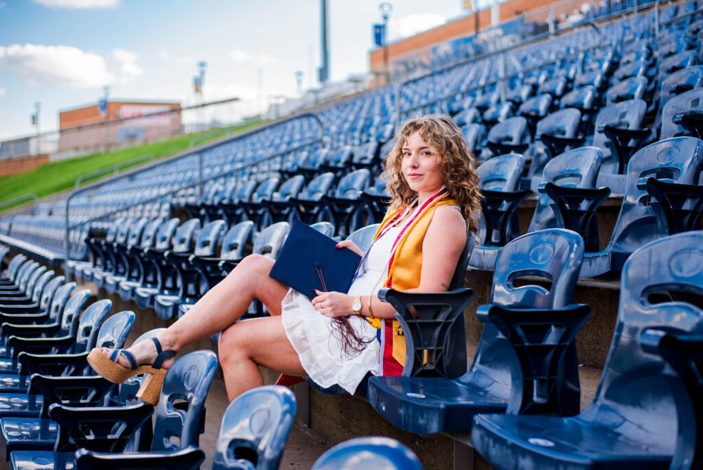 wvubaseballgradphotos11 carrie white photography graduate woman in a white dress with a yellow sash sits in blue stadium seats holding a diploma folder and looking at the camera outside the arena footage