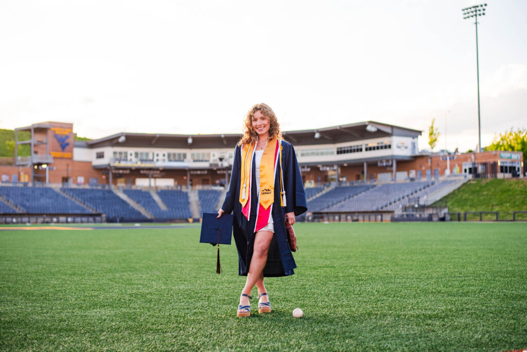 wvubaseballgradphotos28 carrie white photography graduate in navy gown and yellow stole stands on a football field holding a diploma and smiling with stadium seats behind her