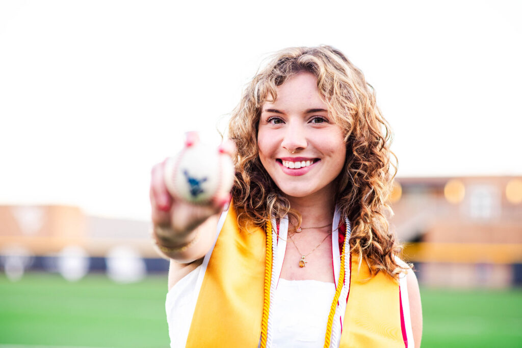 wvubaseballgradphotos41 carrie white photography smiling young woman wearing a yellow graduation stole and cords extending a baseball toward the camera