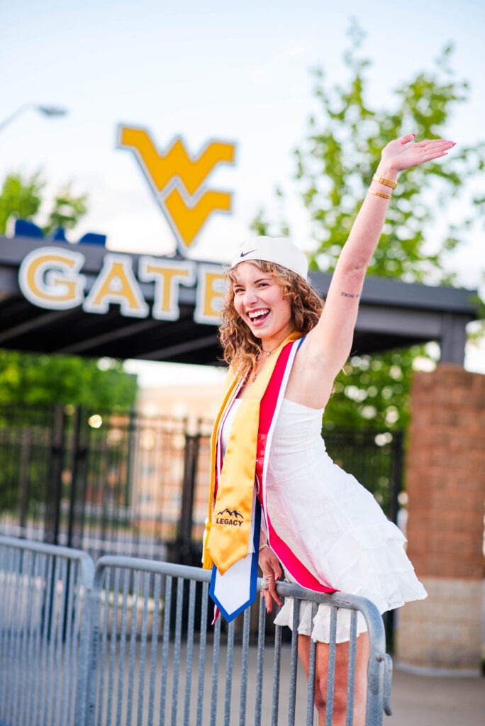 wvubaseballgradphotos42 carrie white photography happy graduate in a white dress and cap raises one arm in celebration with a yellow wv sign in the background