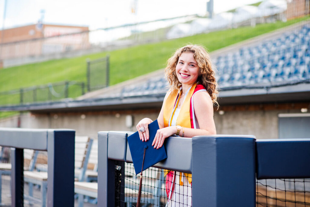 wvubaseballgradphotos52 carrie white photography smiling graduate in a white gown with a yellow stole holding a blue diploma folder near stadium seating