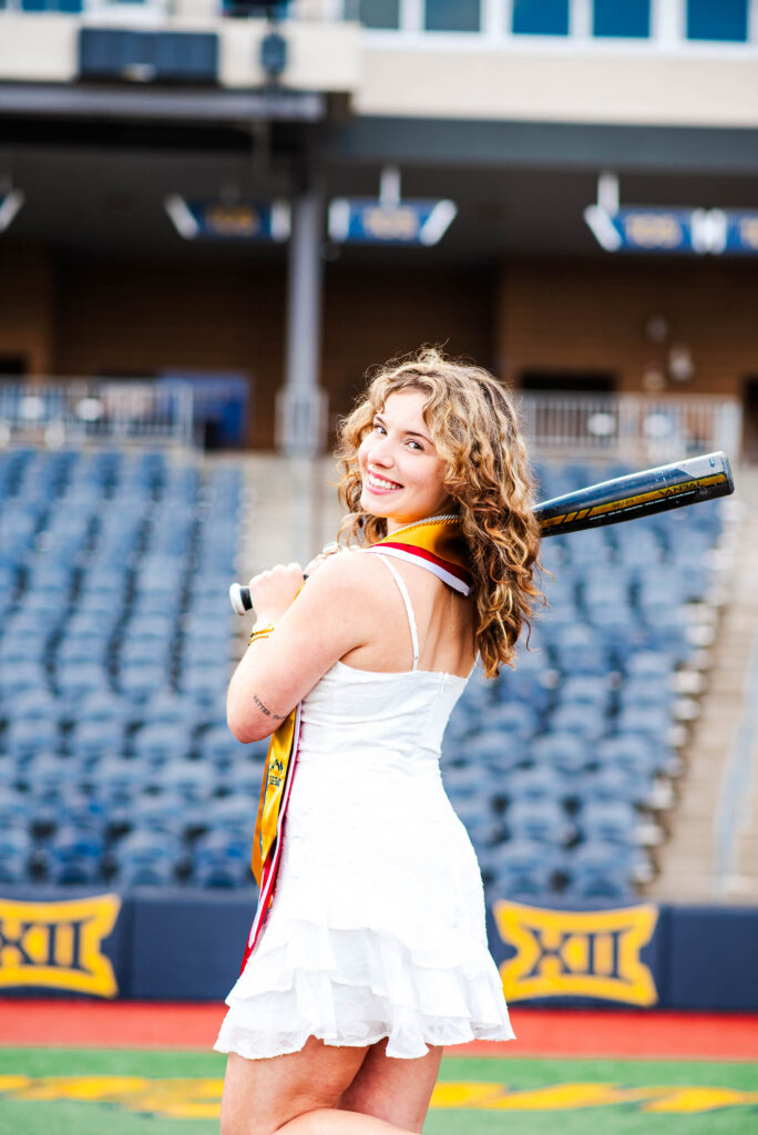 wvubaseballgradphotos54 carrie white photography smiling woman with curly blonde hair in a white dress holds a baseball bat on her shoulder in a stadium