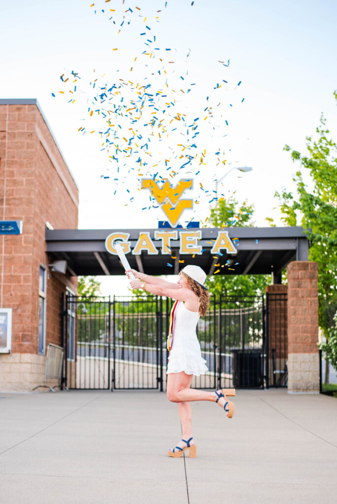 wvubaseballgradphotos57 carrie white photography woman in a white dress and platform sandals throws blue and yellow confetti at a graduation entrance gate a on campus
