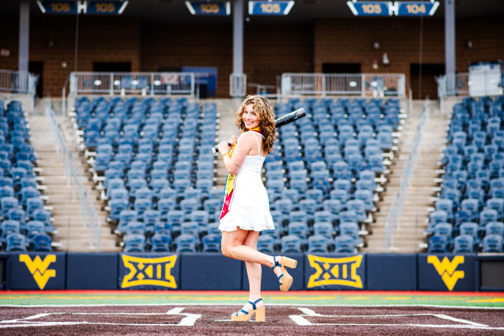 wvubaseballgradphotos61 carrie white photography a female grad in a white dress stands on a stadium field smiling with empty blue seats and yellow banners in the background during baseball graduation pictures