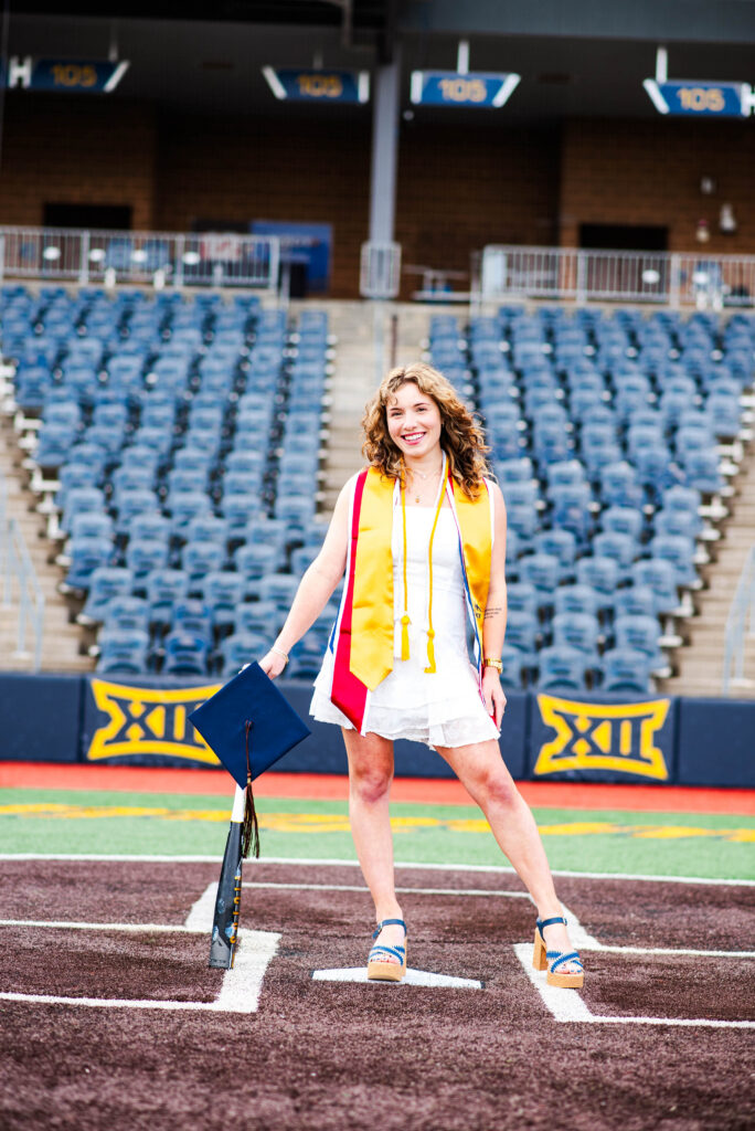 wvubaseballgradphotos68 carrie white photography graduate in a white dress with a yellow stole standing on a stadium field holding a blue graduation cap with tassel visible