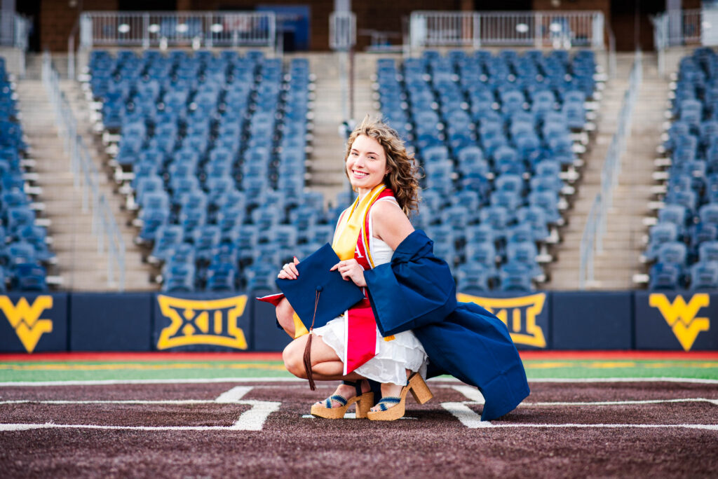 wvubaseballgradphotos82 carrie white photography graduate in a blue cap and gown smiling on a sports field holding her diploma and tassel with empty stadium seats in the background