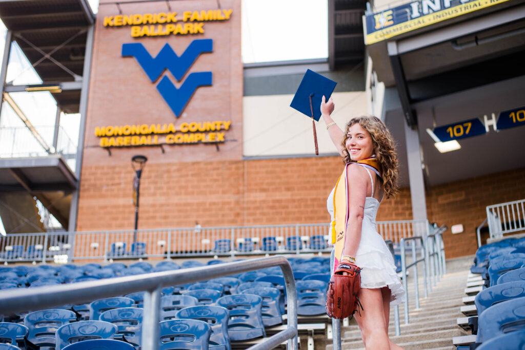 wvubaseballgradphotos83 carrie white photography graduate woman in a white dress raises a blue mortarboard in the air while standing in the stadium seating at kendrick family ballpark