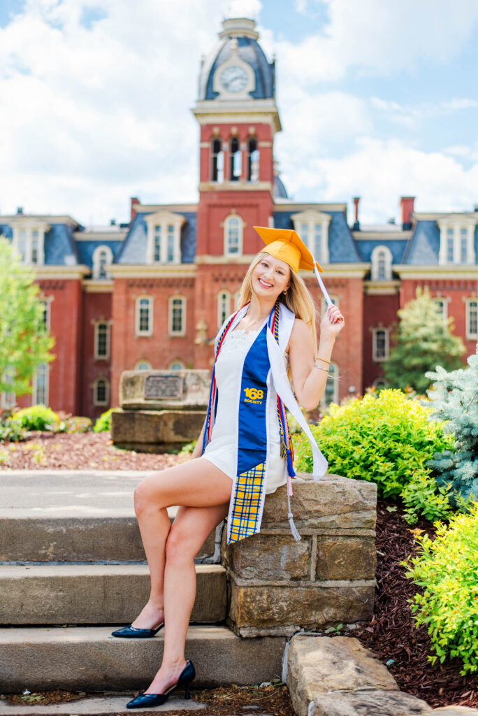 wvucapgowncordsstoles01 carrie white photography graduate in orange cap and blue and white gown sits on campus steps with a red brick university building and clock tower behind her