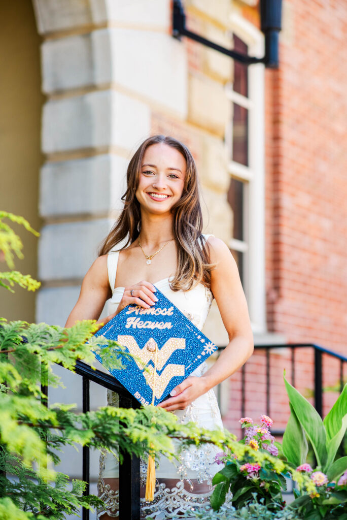smiling young woman in a white dress standing outside near a brick building holding a blue graduation cap decorated with white text