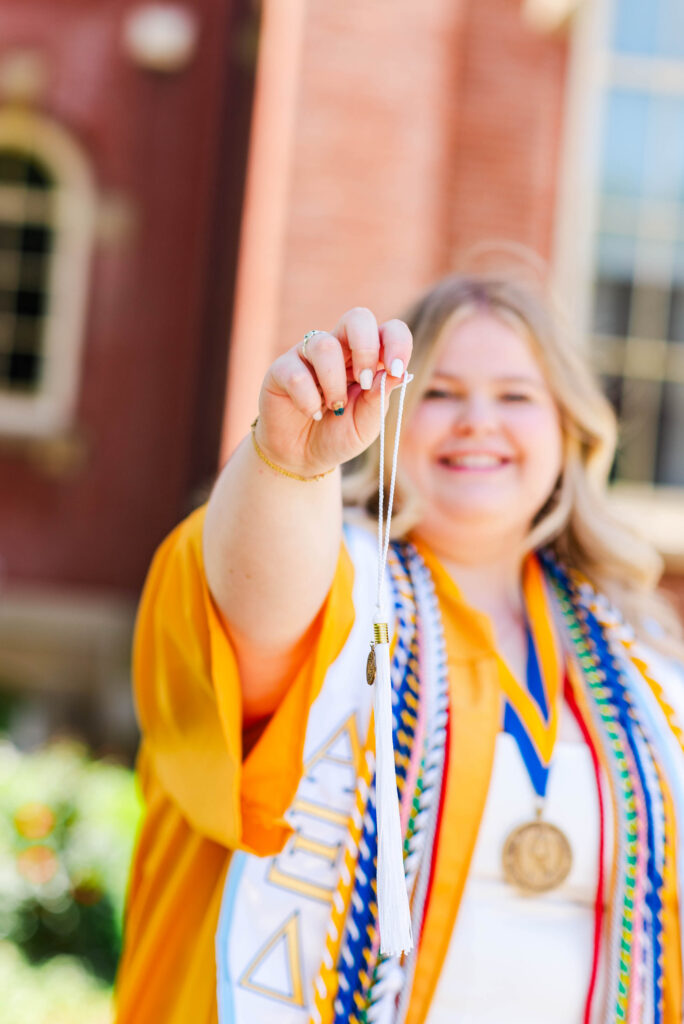 smiling graduate in an orange gown holding up a white tassel and cords with medals around her neck
