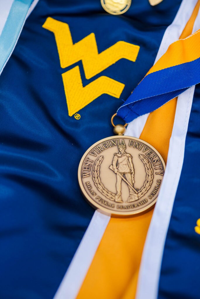 wvucapgowncordsstoles09 carrie white photography gold medal with west virginia university seal resting on blue graduation gown with yellow and blue cordsed ribbons grip the fabric