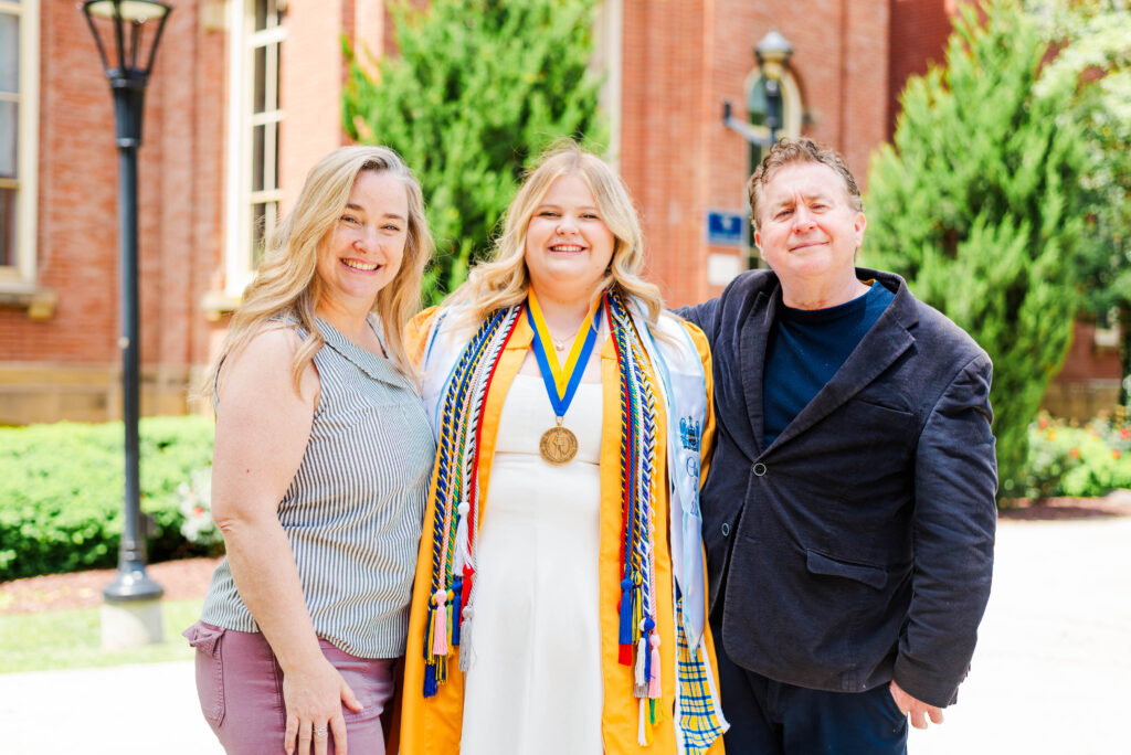 graduate in orange gown with multiple cords and medals flanked by smiling parents outdoors on campus