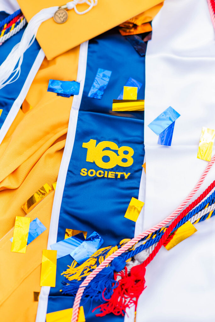 wvucapgowncordsstoles30 carrie white photography close up of a blue and yellow graduation gown with tassels and scattered confetti'168 Society' visible on the fabric.