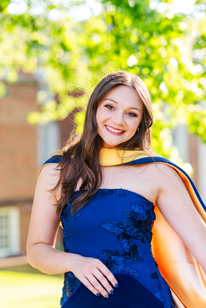 smiling woman in a blue dress with floral embroidery standing outdoors with sunlit green background