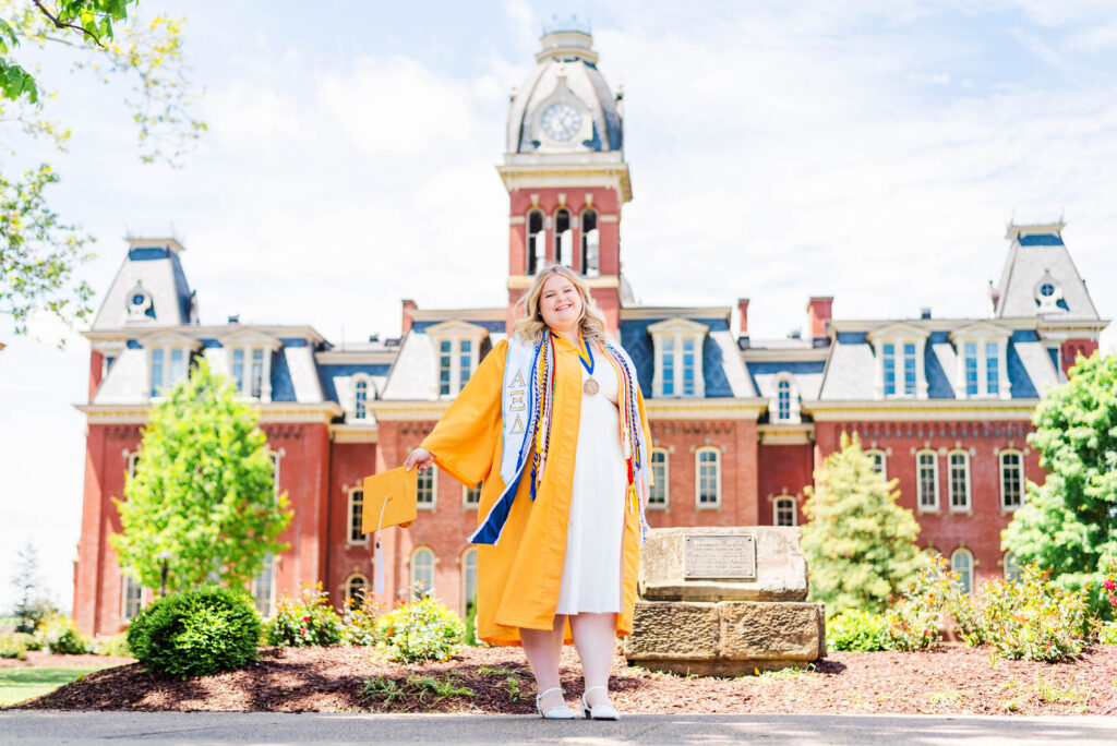 Smiling graduate in a yellow gown with stoles and cords holds a diploma in front of a red-brick campus building with a clock tower on a sunny day during WVU Spring grad photos.