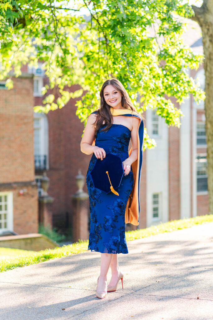 smiling graduate in a blue lace dress holding a blue graduation cap with an orange tassel outdoors among greenery