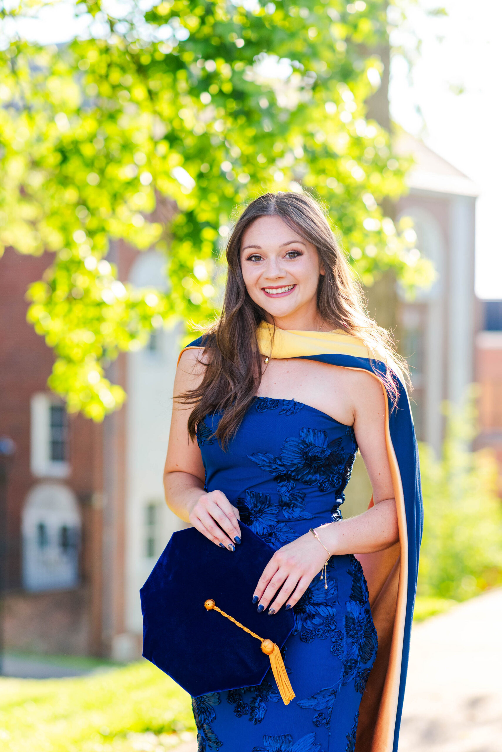 young woman in a dark blue floral dress outdoors holding a navy graduation cap with an orange tassel and smiling at the camera