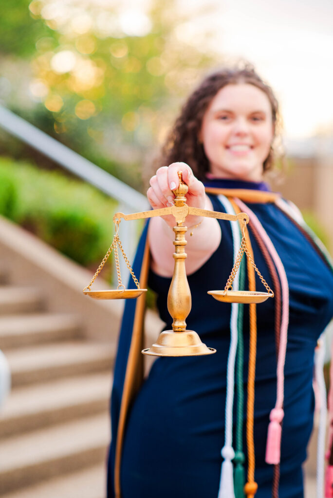 smiling woman holds a gold balance scale toward the camera outdoors symbolizing justice or fairness