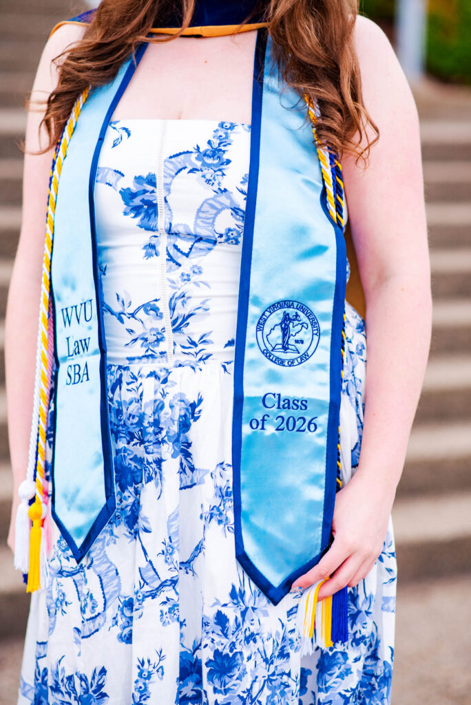 graduate wearing a light blue graduation stole over a blue floral dress wvu law sba and class of  emblem visible on the stole