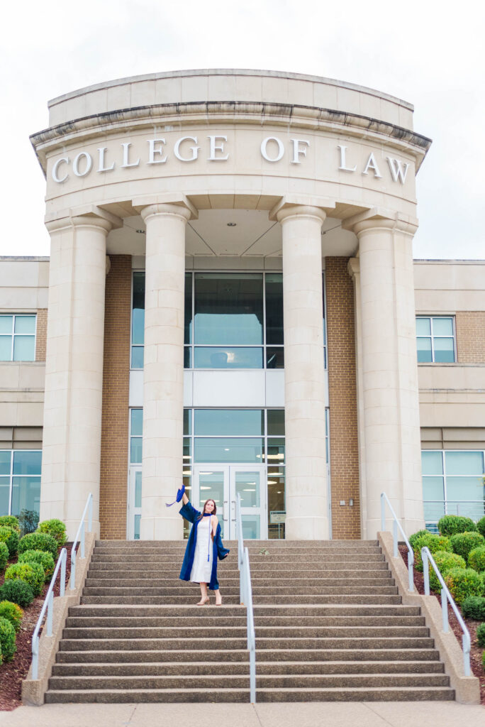 graduate in a blue gown holds a cap on the steps of a campus building with tall columns and'College of Law' signage above