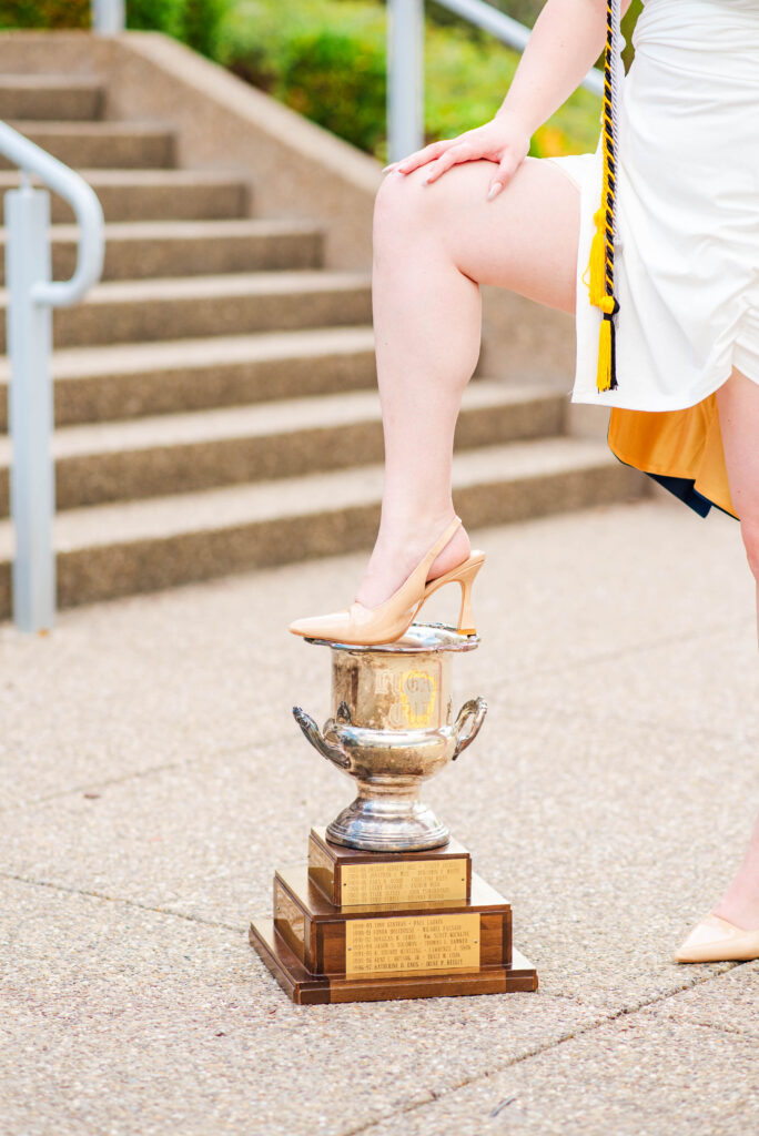person in a white dress with a yellow and black tassel stands on a large silver trophy outdoors one foot resting on the pedestal
