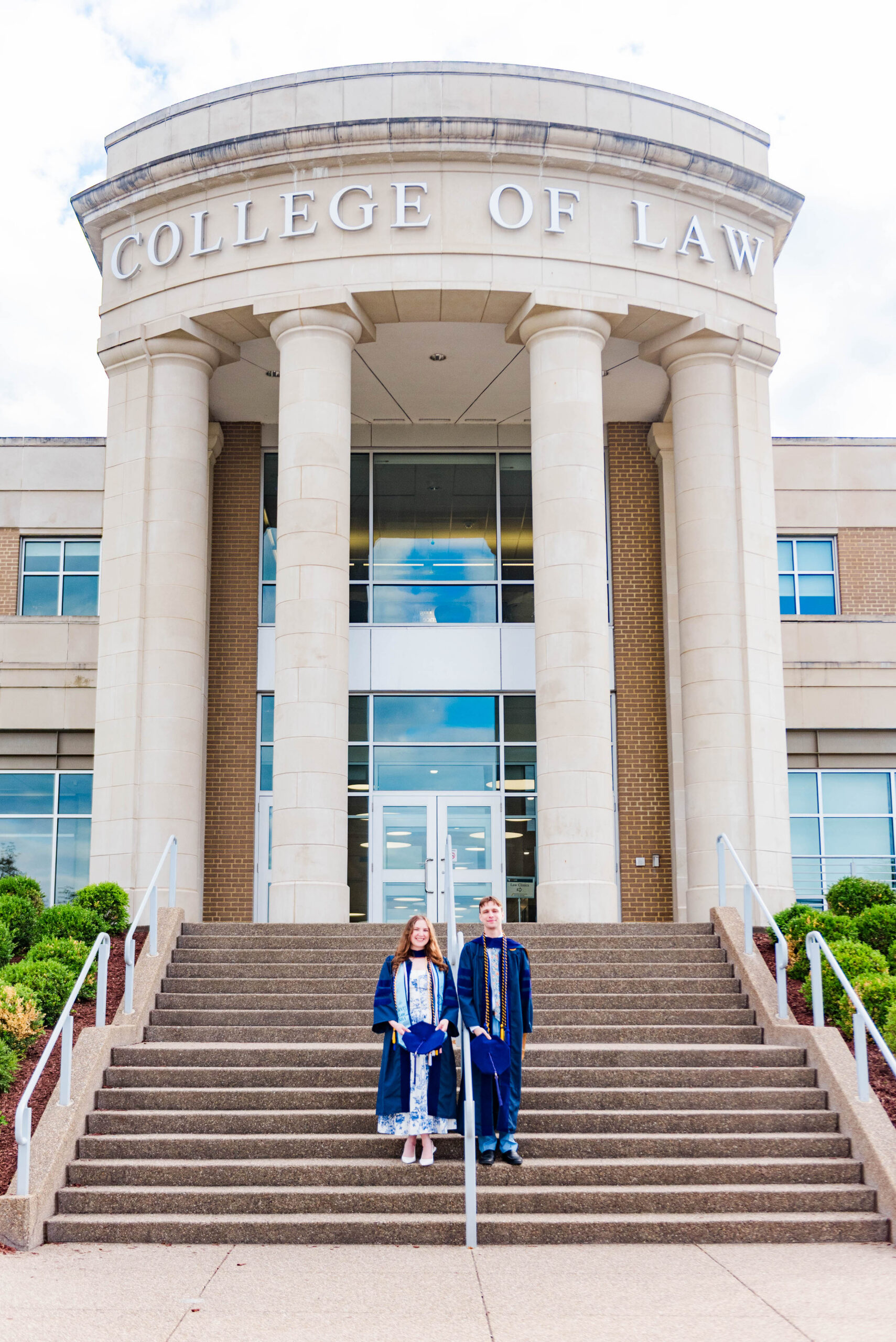 Two graduates in blue gowns pose on the steps of a College of Law building with tall stone pillars and large glass doors.
