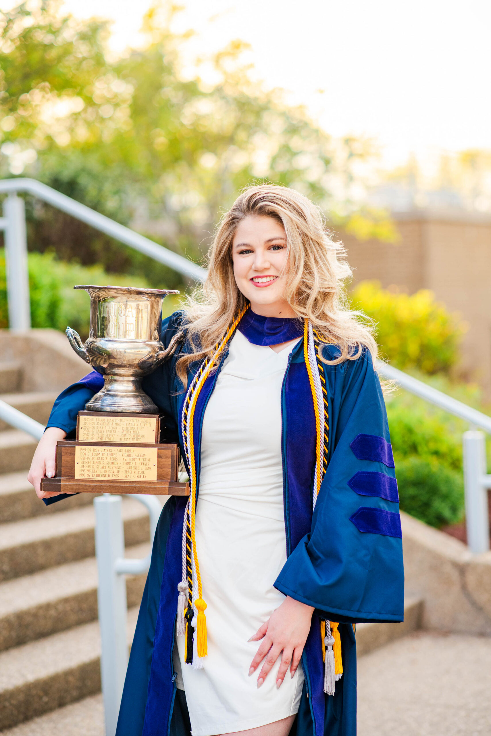 Graduate in a blue gown and white dress poses outdoors, holding a tall trophy with plaques.