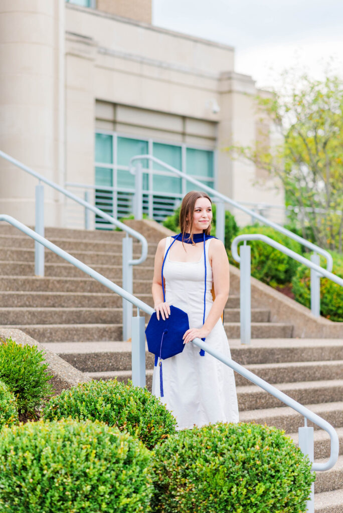 woman in a white dress holding a blue graduation cap on outdoor stairs with a railings and greenery nearby
