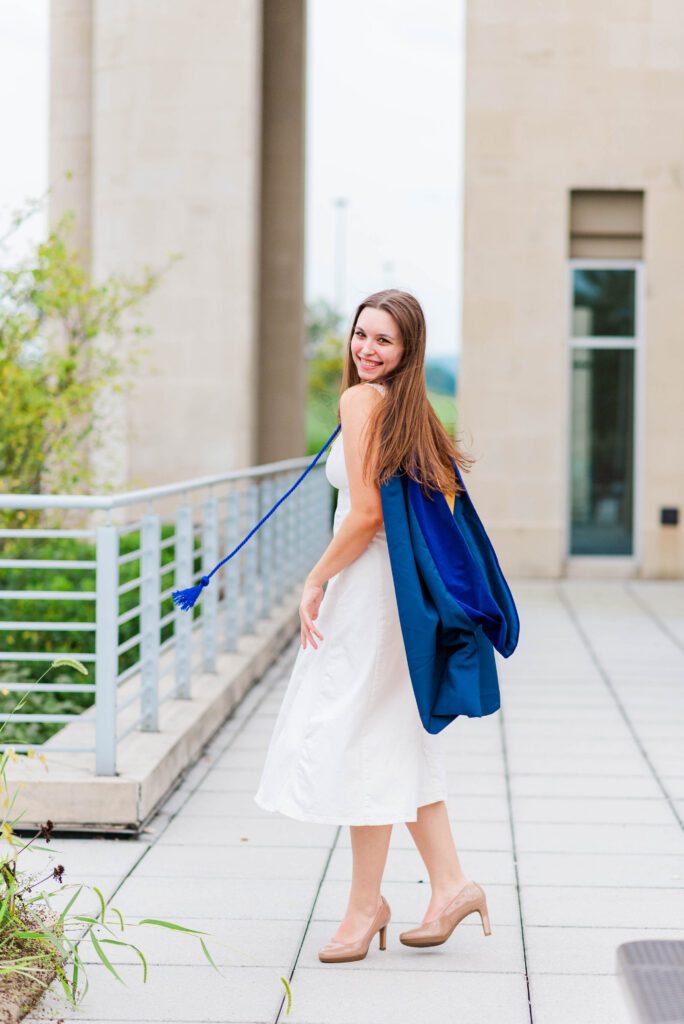 smiling woman in a white dress with a blue cape walks along a tiled path beside a railing outside a beige building