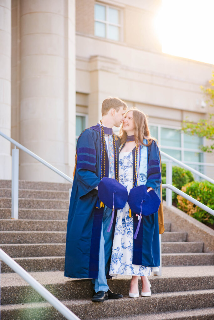 couple in blue graduation gowns holding their blue mortarboards on outdoor steps smiling at each other outside a campus building