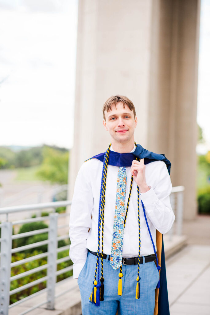 young man in a white shirt with a blue gown and cords posing outdoors beside a tall columned structure alternatively graduate posing with ceremonial cords draped over shoulders outdoors near a railing