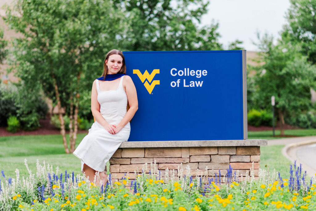 woman in white dress leaning against a blue college of law sign with a yellow logo outdoors among green trees and colorful flowers during a wvu college of law grad photos