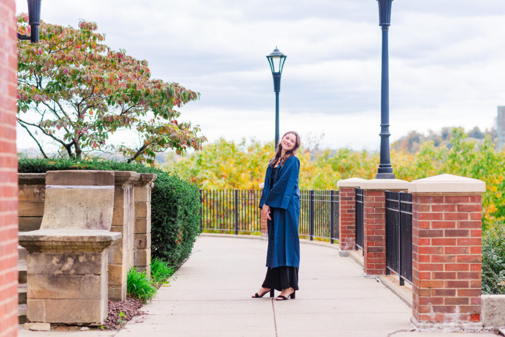 wvudecembergradphotos05 carrie white photography smiling woman in a blue graduation gown posing on a sidewalk with brick pillars and lamp posts autumn trees in the background